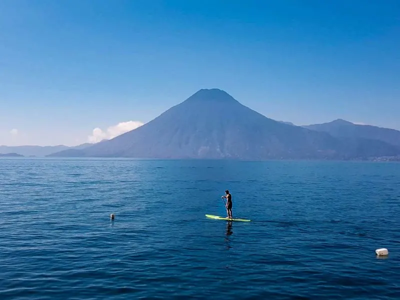 A man paddleboards on the calm waters of Lago Atitlan with a volcanic peak in the background, an activity that you can't miss on a trip to Guatemala