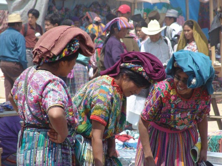 Three women wearing traditional Guatemala huipiles and headpieces converse as they investigate wares at the Mercado San Francisco el Alto, one of the best things to do in Guatemala for tourists