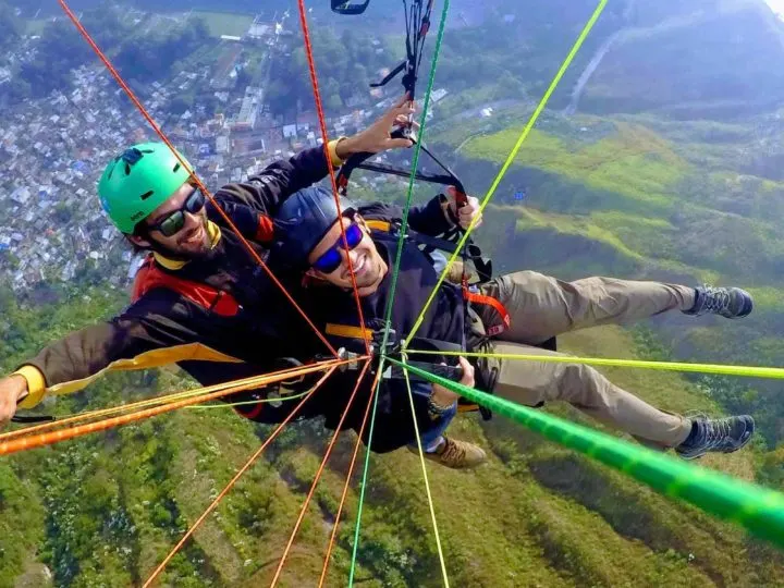 Two men attached to the strings of a paraglide above the hills and a village in Guatemala, one of the most extreme and fun things to do in Gutemala 