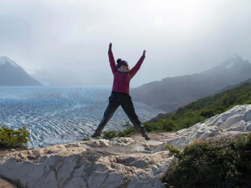 A hiker doing a star jump in front of Glacier Grey while hiking the O Circuit in Torres del Paine National Park, Patagonia
