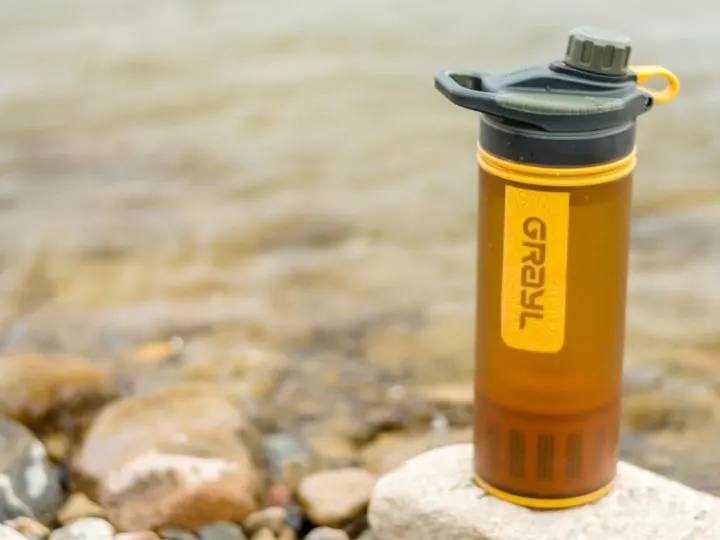 An orange Grayl Geopress travel water filter sits on a rock in front of a river
