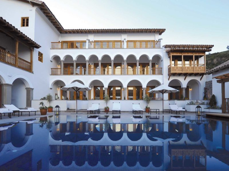 A white hotel with many arched balconies over looks a dark blue swimming pool in Cuzco