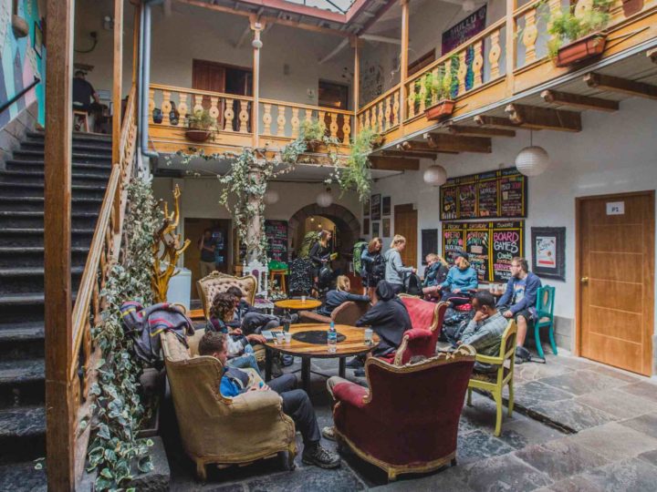 A dozen backpackers chat around a table in the hostel courtyard, they're surrounded by stairs and a wooden balcony.