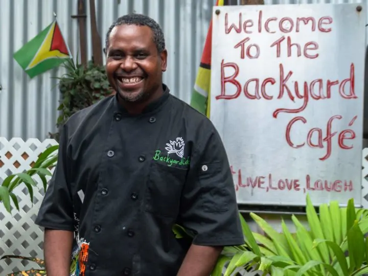 A man smiles in front of a sign for the Backyard Cafe in Guyana.