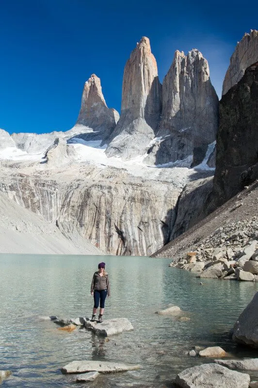 A person stands on a rock in the lake in front of the towers or torres of Torres del Paine National Park, a viewpoint accessible on a day hike in the park