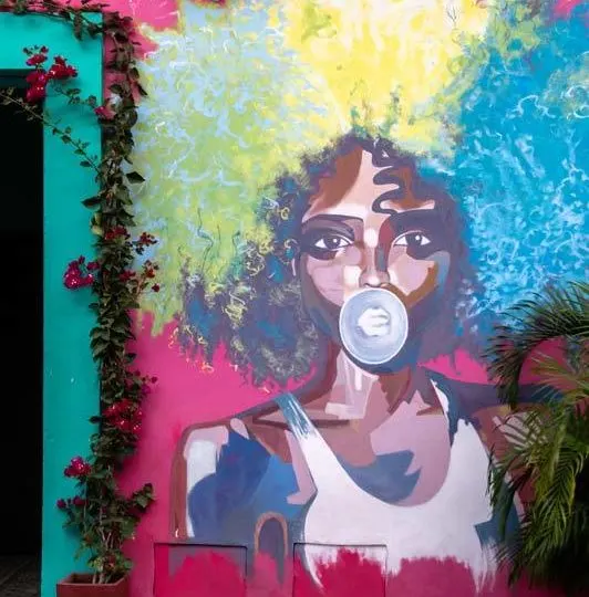A Black afro-Caribbean woman with a rainbow-coloured afro blows bubble gum on a piece of street art in Cartagena, Colombia 