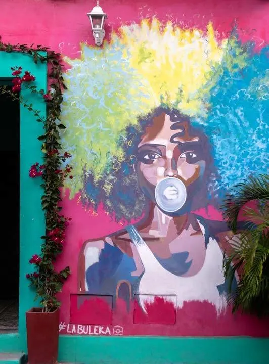 A Black afro-Caribbean woman with a rainbow-coloured afro blows bubble gum on a piece of street art in Cartagena, Colombia