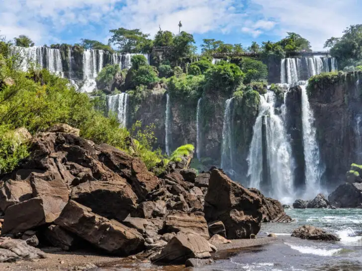 The Iguazu Falls in Argentina as they cascade into the pool below them. The image shows rocks in the foreground, with multiple waterfalls in the background.