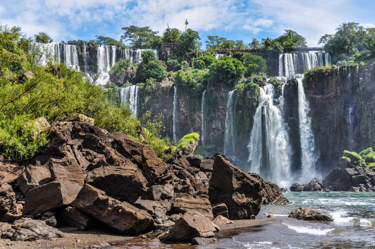 The Iguazu Falls in Argentina as they cascade into the pool below them. The image shows rocks in the foreground, with multiple waterfalls in the background.